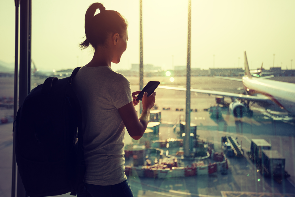 Viajante olhando o celular em frente à janela do aeroporto, com aviões ao fundo