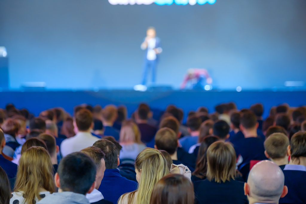 Palestrante no palco de um evento de tecnologia falando para uma plateia grande em um auditório com iluminação azul