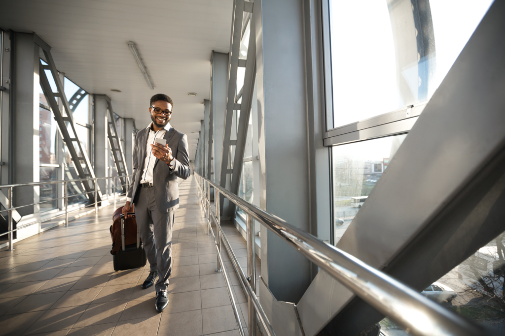 Homem sorrindo caminhando com mala no terminal do aeroporto enquanto usa aplicativo no celular